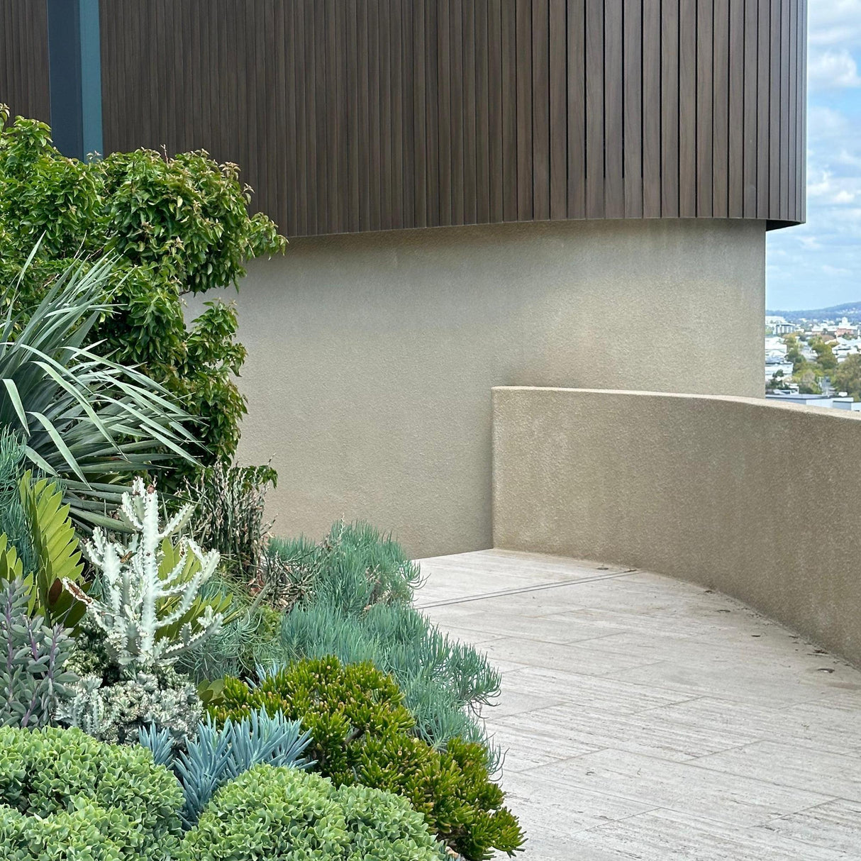 Experience outdoor space transformation with our Garden Design Consultation, featuring a curved concrete path, lush green plants, and a modern wood-paneled building wall set under a cloudy sky.-Nursery Near Me