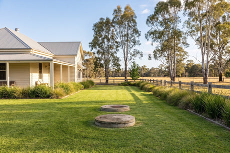 Sunny mown lawn over a septic drain field beside a farmhouse, with tank lids, a low grass border and gum trees well back