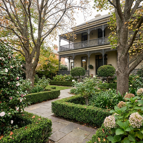 A stone path winds through a lush, manicured garden toward a large house with a balcony and ornate railings.