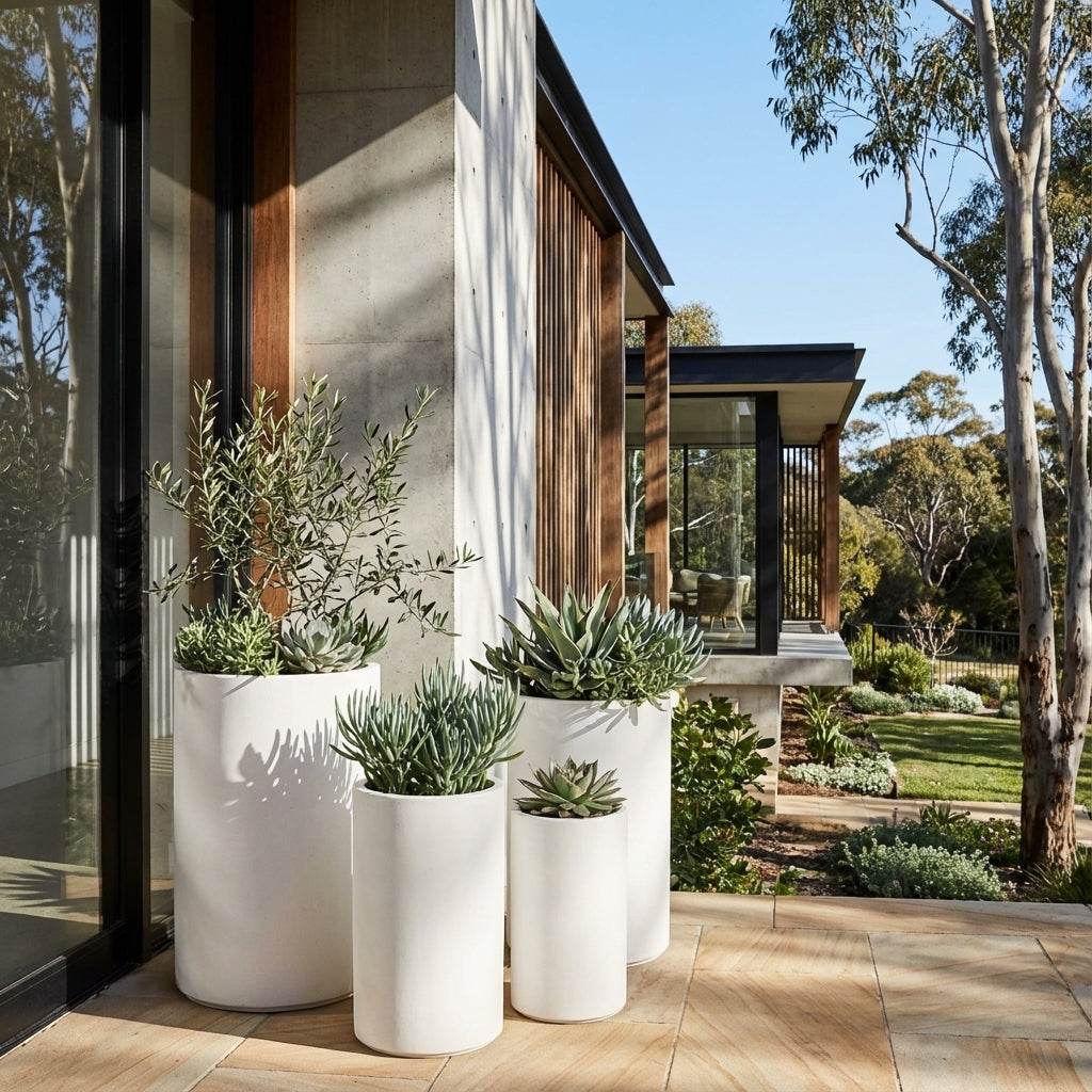Four White Harper Cylinder Pots in various sizes, made from weather-resistant fibreglass, showcase succulents and greenery to add modern charm beside the glass-walled house.