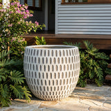 The Grey White Grid X Large Cement Pot (700mm x 590mm) features a modern grid texture, making it a striking decorative planter among lush green plants on a stone patio.