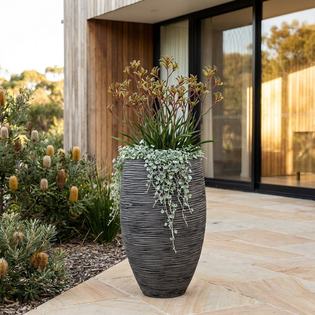 The Rib Outdoor Plant Pot, available in various sizes, features a tall, textured design with a natural stone look and double-walled insulation—perfect for showcasing green and red foliage on your patio by a modern house and garden.