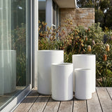 Four White Harper Cylinder Pots in various sizes are made from lightweight fiberglass. They sit on a deck by a glass door, with greenery behind, adding modern, weather-resistant style to any patio or outdoor space.