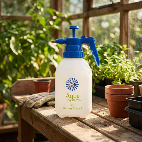 The Aqua Systems 2L Garden Pressure Sprayer with a blue handle is placed on a wooden table next to potted plants in a greenhouse, ready for plant care.