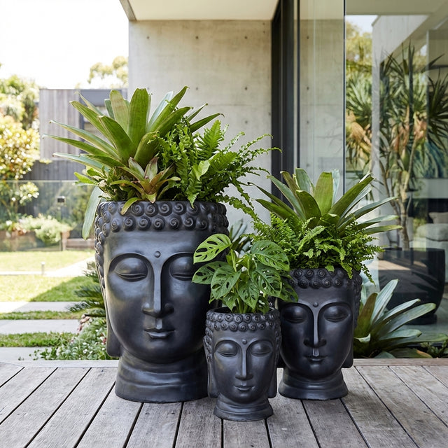 Three Black Balinese Head Planters in various sizes, filled with lush green plants, sit on a wooden deck outside a modern house, adding a striking artistic touch to the outdoor space.