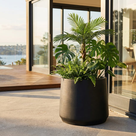 A Black Fibre Stone Round Planter (340 x 290mm) filled with lush tropical plants sits on a modern patio beside glass doors, overlooking the water.
