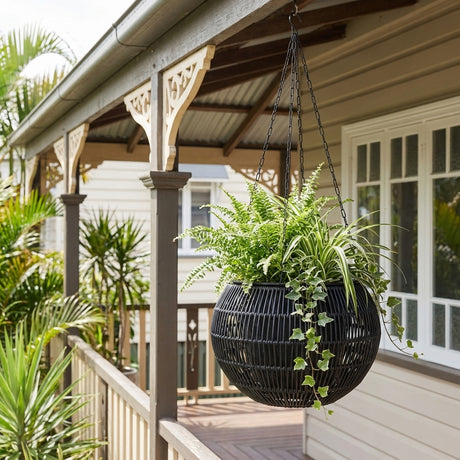 The Black Rattan Hanging Basket - 320mm x 220mm, filled with lush ferns and ivy, hangs beautifully on a porch amid greenery and wooden railings—an ideal outdoor hanging basket for any stylish home.