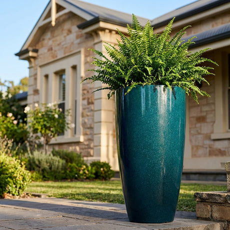 A large green fern is displayed in a Blue LA Bullet Pot - 310mm x 530mm in front of a stone house, adding a stylish, contemporary touch that's perfect for indoor or outdoor arrangements.