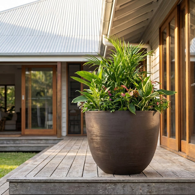 A White Maxim Drum Pot filled with green leafy plants sits on a wooden porch outside a modern house with glass doors. Various sizes available.