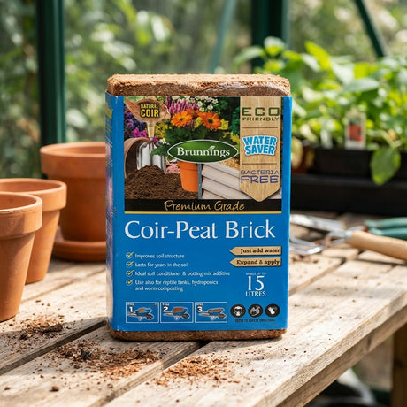 A Brunnings 15L Coir Peat Brick rests on a potting bench in a greenhouse, surrounded by pots and gardening tools, ready to be used as an eco-friendly soil conditioner with excellent water retention.