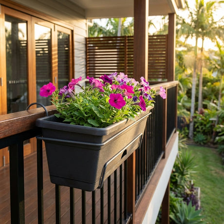 A Charcoal Balcony Planter - 500mm filled with pink and purple flowers hangs on a balcony railing, catching the sunlight.