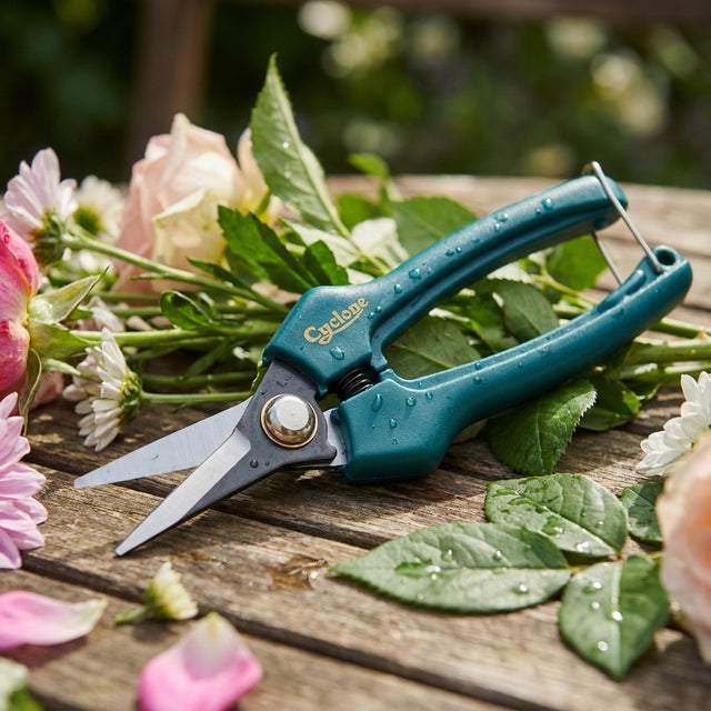 Cyclone Pruner Floral Snips with water droplets rest on a wooden table among freshly cut flowers and leaves, ready for precise trimming tasks.