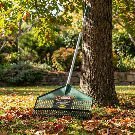 A Cyclone Super Garden Rake leans against a tree on a lawn blanketed with fallen autumn leaves.