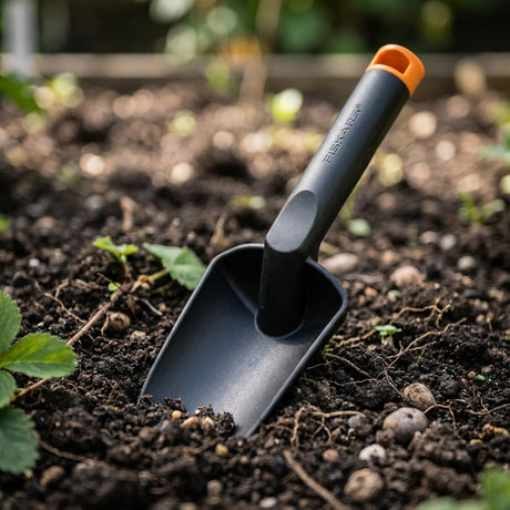 A Fiskars Nyglass Trowel with a black head and orange handle is shown in soil among small plants—an ergonomic tool ideal for precise gardening tasks.