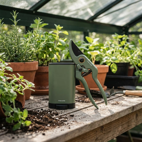Fiskars Plant Care Pruner With Holder in green, placed on a wooden table in a greenhouse surrounded by potted herbs and plants.