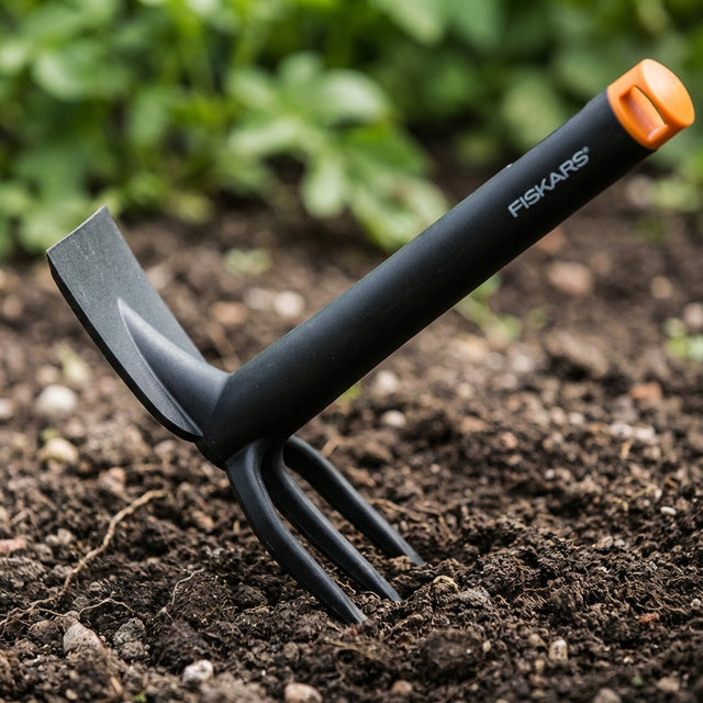 A black Fiskars Planter Hoe with an orange tip, an ergonomic garden tool ideal for planting and weeding, is stuck into the soil in a garden.
