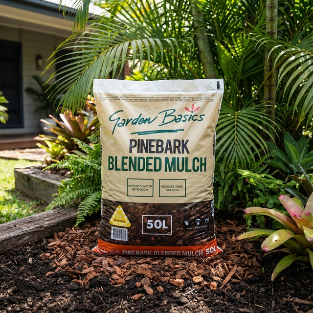 A bag of Garden Basics 50L Pine Bark Mulch sits in a garden with plants and a house in the background, providing great water retention and weed control for lush, healthy landscapes.