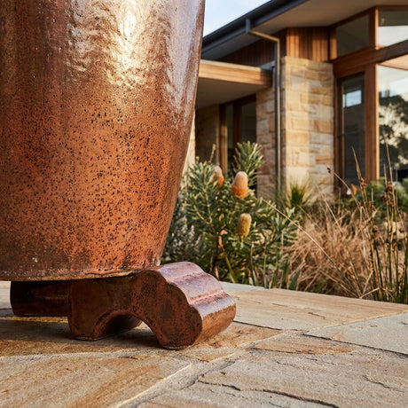A large copper planter sits outdoors on a Glazed Pot Foot, with a modern house and native plants in the background.
