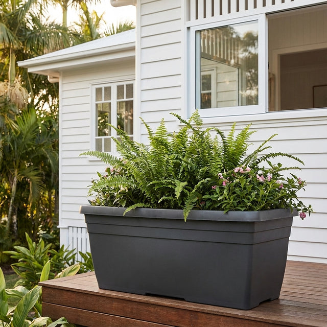 A Graphite Villa Trough Pot (various sizes available) filled with ferns and flowers sits on a wooden deck outside a white house, adding charm to the outdoor space with its stylish plant trough design.