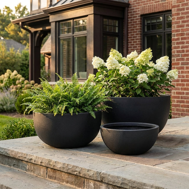 Three Grey Harper Bowl planters in different sizes, filled with green ferns and white flowers, sit on stone steps outside a brick house.