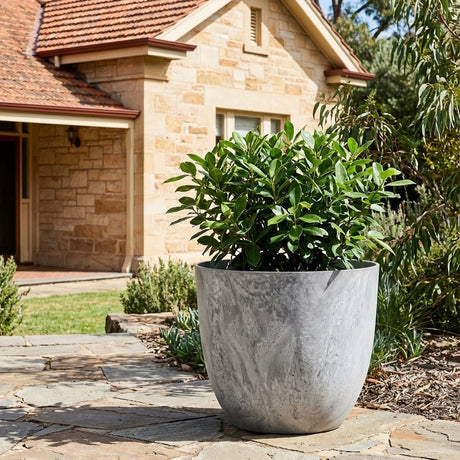 A lush green plant is displayed in the Grey Stone Art Egg Pot (available in various sizes) on a stone patio outside a tan brick house with a red-tiled roof.