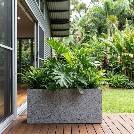 A Grey Terrazzo Trough (various sizes available) filled with green tropical plants sits on a wooden deck by a modern house and lush garden, seamlessly blending style and nature.