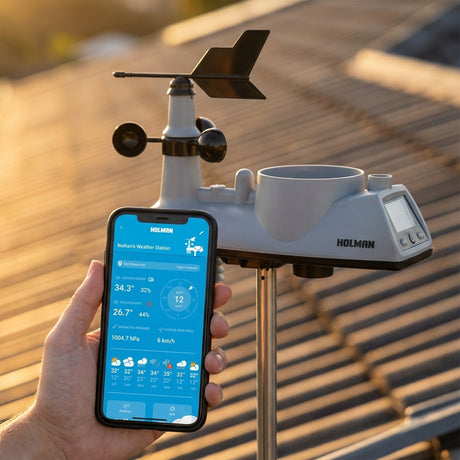 A hand holds a smartphone showing weather data from the Holman Helios Wi-Fi Weather Station next to a rooftop setup at sunset.