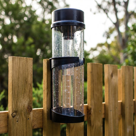 The Holman Professional Rain Gauge Cylinder, with clear measurement markings and outdoor durability, offers precise rainfall measurement when mounted on a fence amidst greenery.