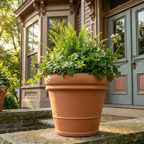 An Italian Terracotta Rolled Rim Pot - 500mm with ferns and ivy sits on a stone porch near a Victorian-style house.