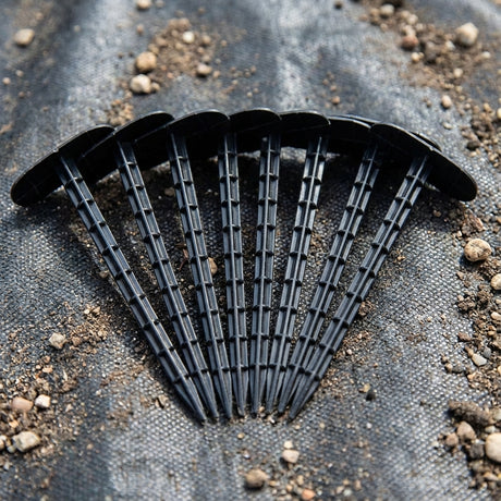 Ten Lotus Collection 14cm Weedmat Pins from the 10 Pack are arranged in a fan shape on soil and landscape fabric.