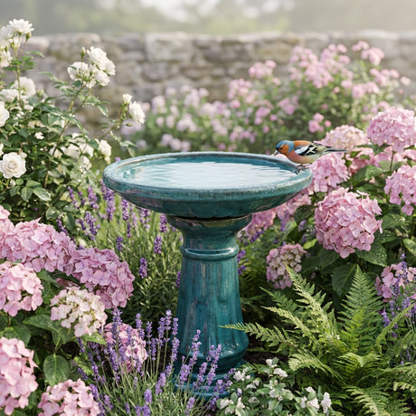 A bird drinks from the Northcote Pottery 52 x 52 x 60cm Forest Green Primo Miami Bird Bath, surrounded by pink and white flowers, creating a charming garden feature.