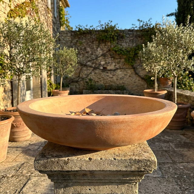 The Northcote Pottery Terracotta Cottaseal Bird Bath Bowl (47 x 11cm) sits on a stone pedestal in a sunny courtyard with potted plants and stone walls.