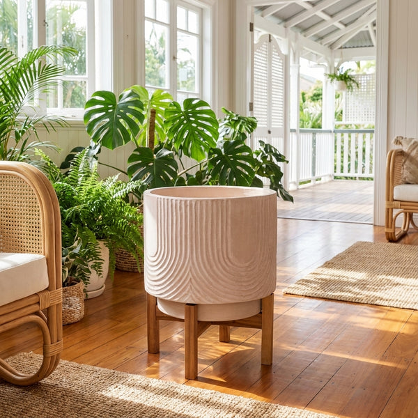 A Pink Indoor Ceramic Pot With Stand (340mm x 430mm) sits in a bright sunroom, surrounded by green plants and rattan furniture.