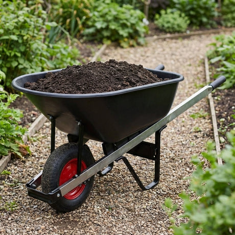 A Gorilla Carts Poly Dump Cart - 115L, filled with soil, is positioned on a gravel garden path amid lush green plants.