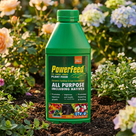A green bottle of PowerFeed Concentrate rests on garden soil among blooming flowers, demonstrating how this liquid plant food boosts soil health.