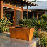 Rectangle Taper Planter in a rust-look finish is displayed on a patio with vibrant greenery, complementing the modern wood-framed house. Various sizes available.