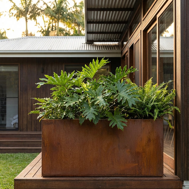 A Rectangle Taper Planter with lush green tropical plants sits on a wooden deck beside a modern house. Various sizes available.
