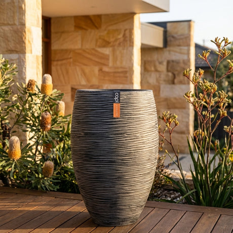 Large grey Rib Outdoor Plant Pot (various sizes available) on wooden deck with native plants, stone building in background.