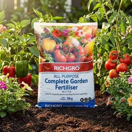 A bag of Richgro All Purpose Complete Garden Fertiliser rests among tomato, pepper, and flowering plants in a garden.