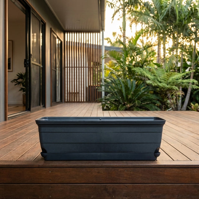 The Roman Self Watering Trough Planter - 600mm, a sleek black and lightweight rectangular planter, sits on a wooden deck with lush plants in the background.