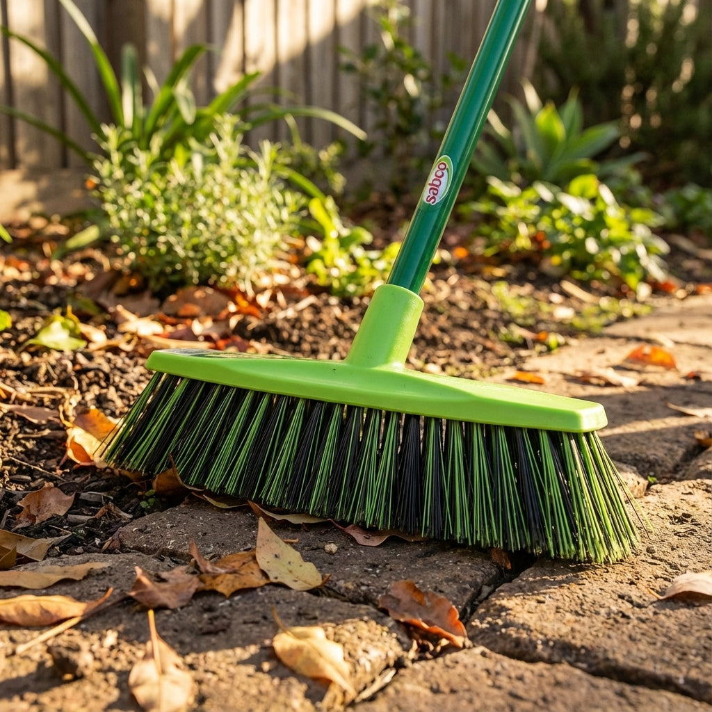 The Sabco Premium Yard Broom, featuring crimped bristles, efficiently sweeps dry leaves from a stone patio in a sunlit garden.