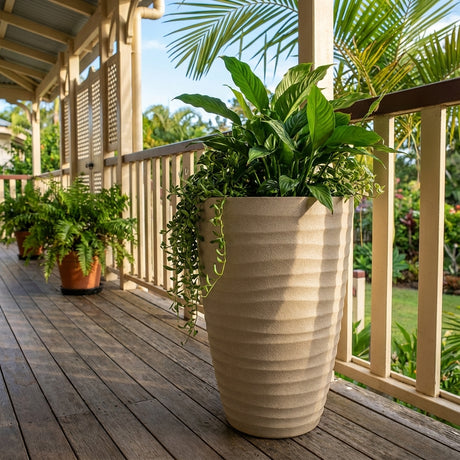 The Sandstone Tall Wavy Planter (406mm x 550mm) adds style to any area—ideal for indoor or outdoor use, with green leafy plants on a wooden porch and surrounded by lush garden foliage.