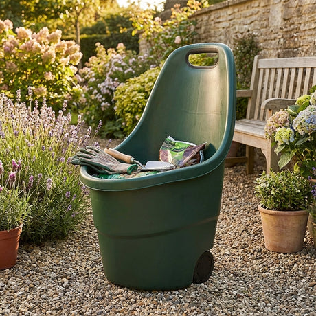 The Saxon Portable Garden Cart - 48 Litres, filled with tools and soil bags, rests on gravel beside a wooden bench and blooming plants in a sunny garden.