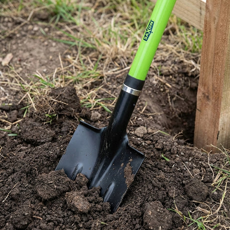 The Saxon Post Hole Long Fibreglass Handle Shovel digs into soil beside a wooden structure in the garden.