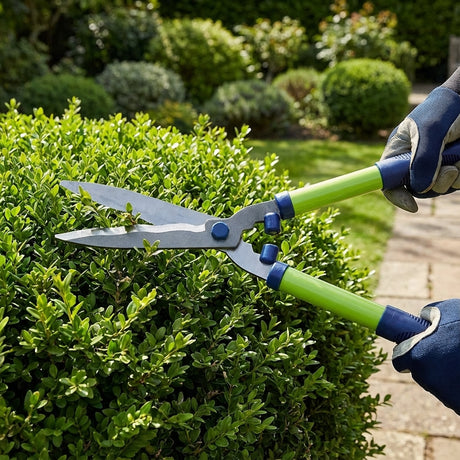 Gloved hands trimming a green bush with the Saxon Wavy Hedge Shear - 550mm in a sunny garden.