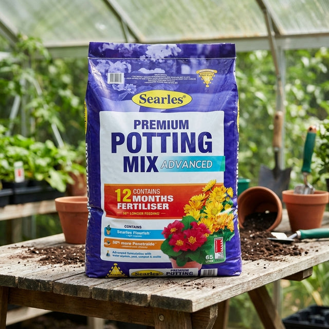 A 65L bag of Searles Premium Advanced Potting Mix rests on a potting bench in a greenhouse, surrounded by healthy, thriving plants.