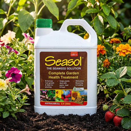 A Seasol 2.4L Complete Garden Health Treatment bottle, a natural seaweed plant tonic, sits among colorful flowers and strawberries in a vibrant garden.