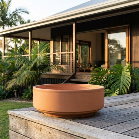 A Terracotta Italian Cylinder Bowl, available in various sizes, rests on a wooden table outside a house with tropical plants and a porch, creating Mediterranean garden decor.