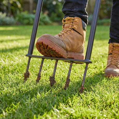 A person uses the Trojan All Steel Spiked Aerator to aerate their grass, while wearing tan work boots on a sunny day.