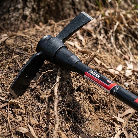 The Trojan Fibreglass Handle Mattock With Cutter End - 2kg sits on dry soil among roots and dried leaves, highlighting its durable black fiberglass handle and robust cutter end.